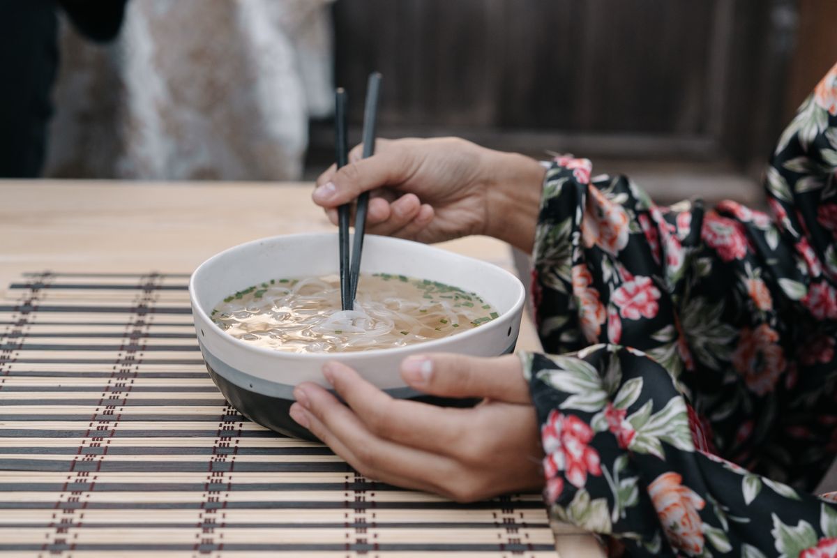 A woman wearing a floral kimono uses chopsticks to eat noodles from a bowl, showcasing traditional dining.