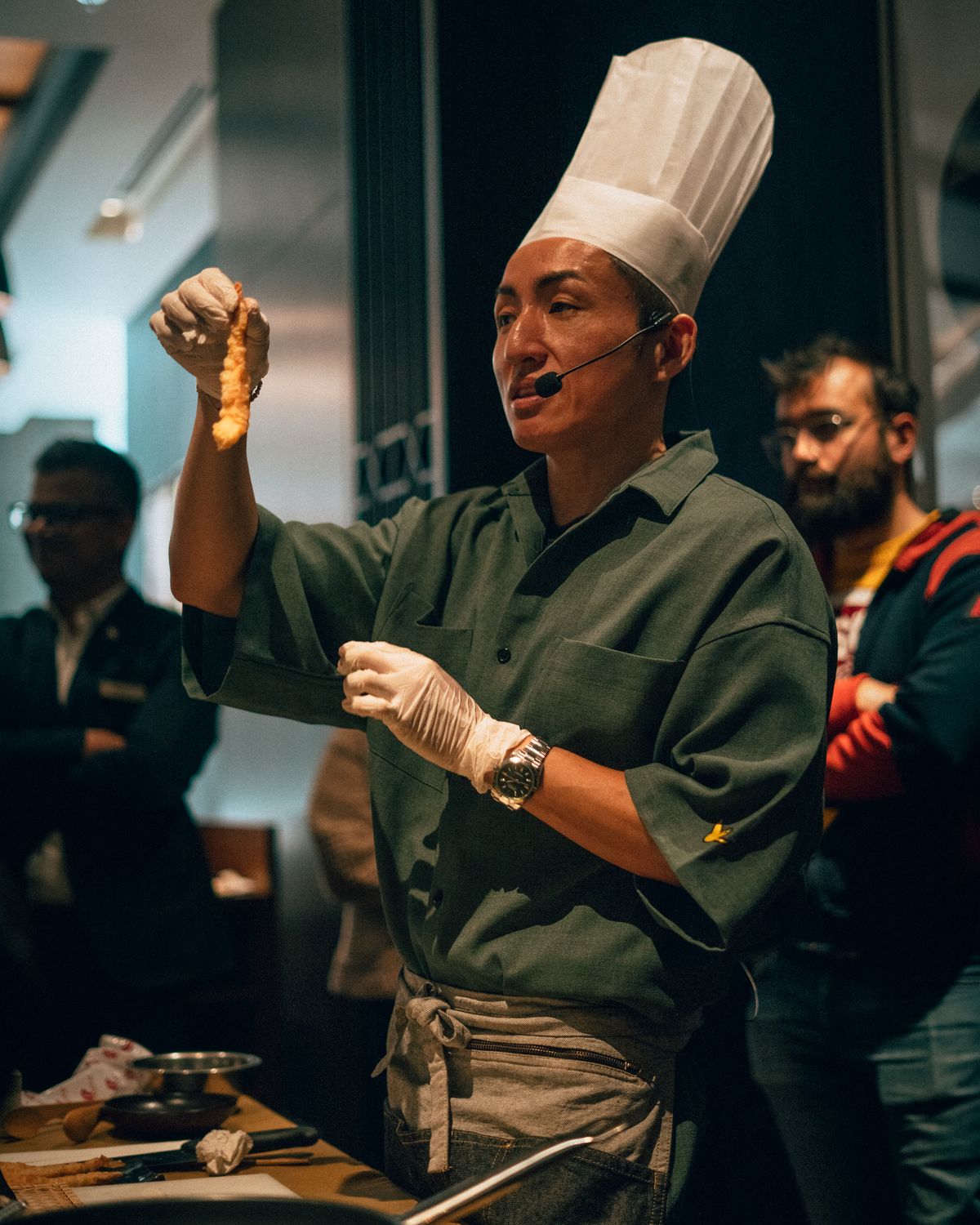 Chef wearing a hat and gloves demonstrates cooking techniques to an audience at an indoor event.