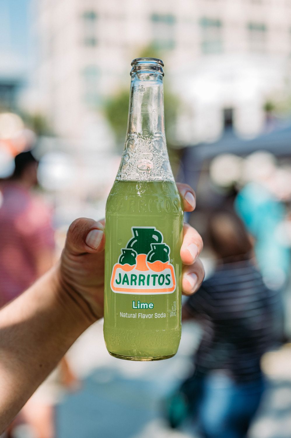Close-up of a hand holding a Lime Jarritos soda bottle in an outdoor setting.