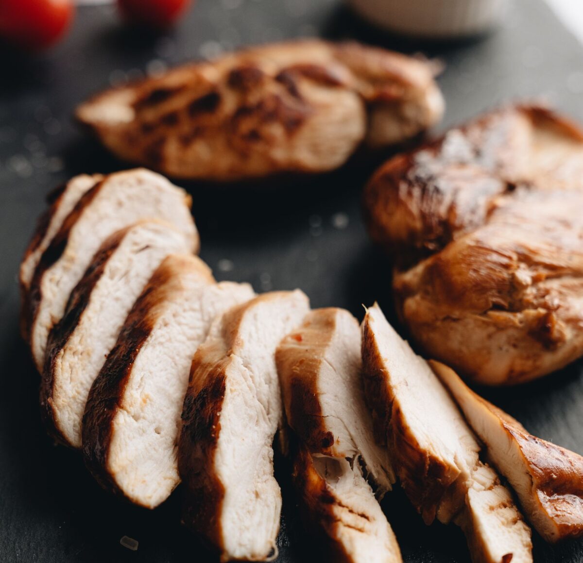 Close-up of grilled chicken breasts sliced on slate with tomato garnish.