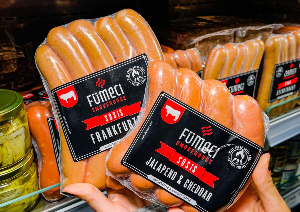 Close-up of hands holding packaged sausages in a grocery store refrigerator section.