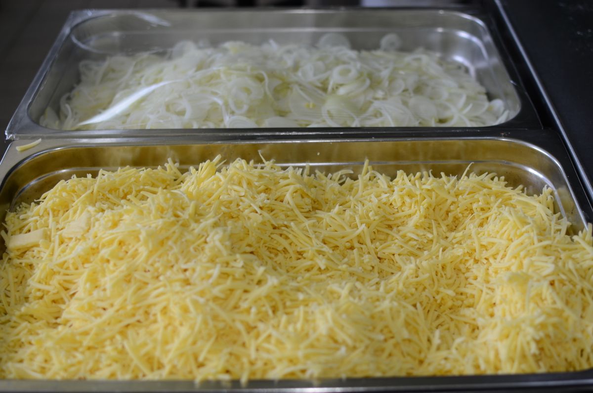 Before Chopped Cheese Went Viral, New York’s Bodegas Already Knew It Was a Star 2 Close-up of shredded cheese and sliced onions in metal trays, perfect for cooking or catering setups.