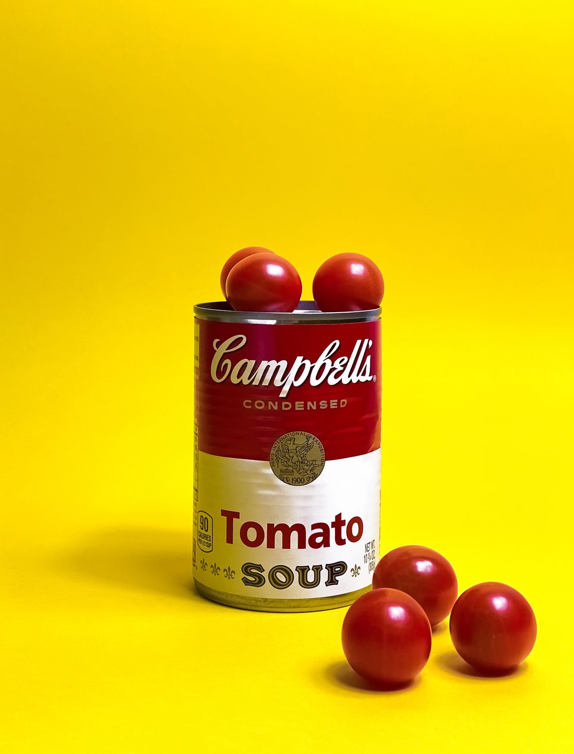 Colorful image of Campbell's tomato soup can surrounded by cherry tomatoes on yellow background.
