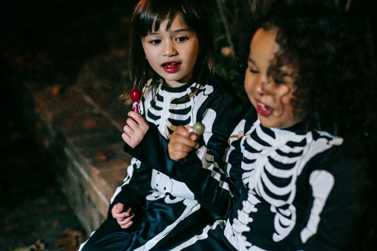 Crop cheerful multiracial girls wearing skeleton costumes with sweet stick candies sitting on brick border in autumn park during Halloween celebration