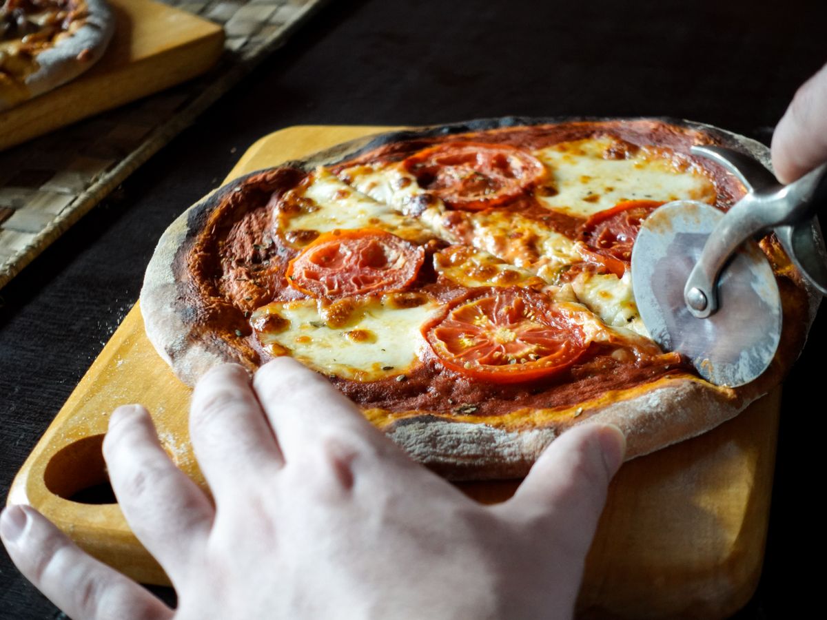Freshly baked homemade pizza being sliced on a wooden board, topped with tomatoes and mozzarella.