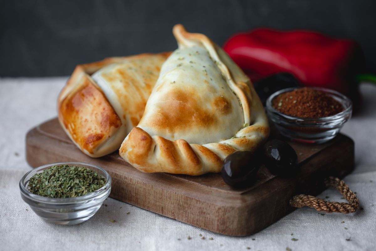 Golden-brown empanadas on wooden board with herbs and spices.