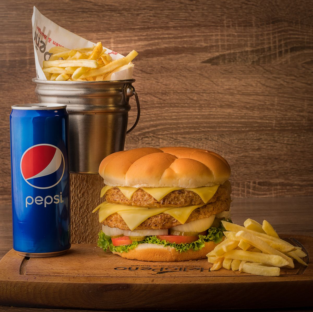 Mouthwatering double cheeseburger with fries and a can of Pepsi on a wooden board.