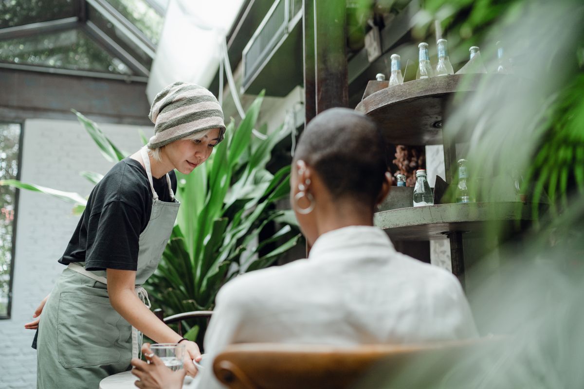 Positive young Asian waitress in uniform smiling and serving delicious food to female customer in modern cozy restaurant full of lush plants