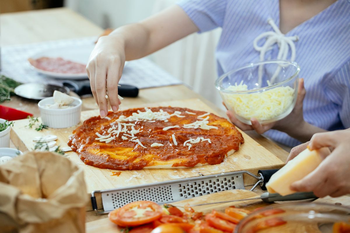 Side view of crop unrecognizable women sprinkling cheese from bowl on yummy homemade pizza while cooking together in kitchen