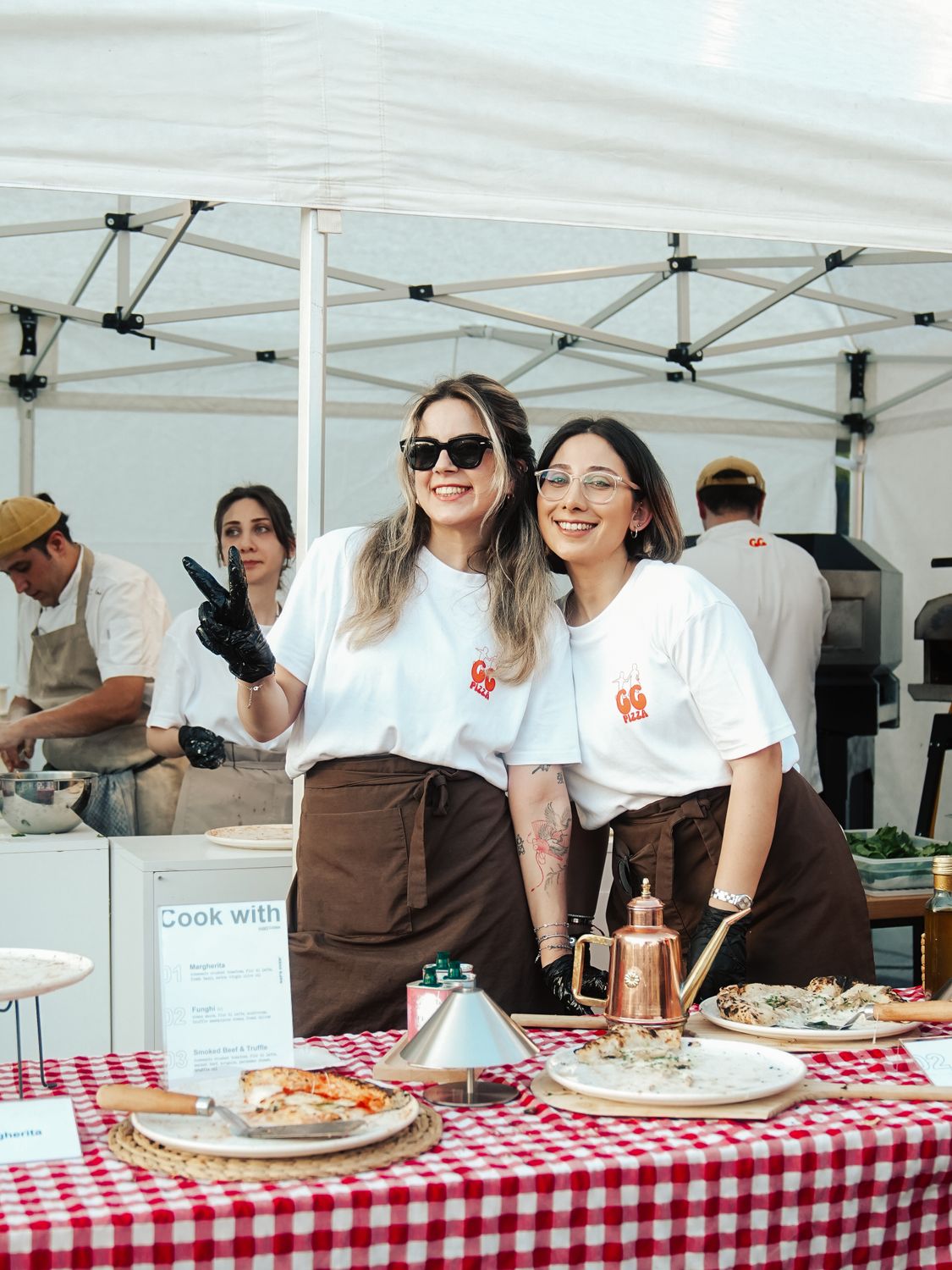 Two smiling women at a food stall during an outdoor market event, promoting a friendly atmosphere.